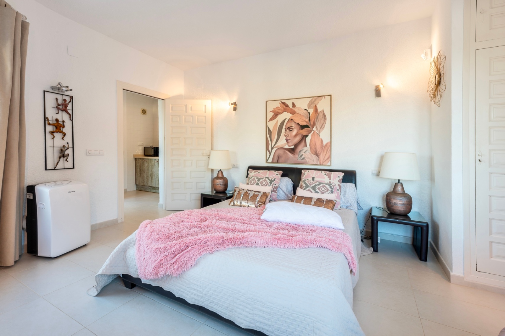Bedroom featuring a double bed with pink accents and built-in white wardrobes. The space includes light tiled flooring, wall-mounted sconces, and a portable air conditioning unit.