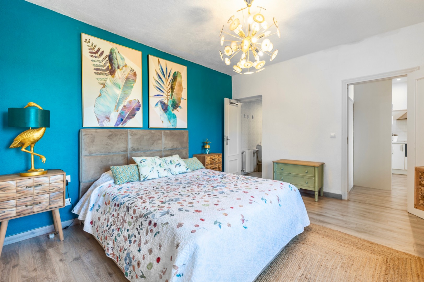 Bedroom featuring a turquoise accent wall, grey upholstered headboard, and botanical artwork. It includes a gold bird-shaped lamp, modern chandelier, and light wood flooring.