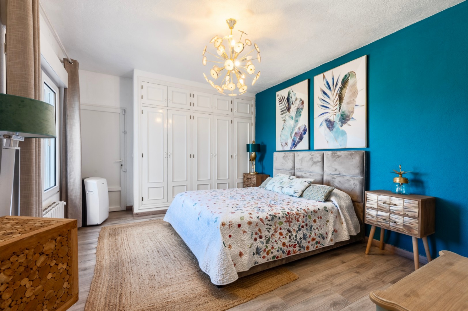 Bedroom featuring a turquoise accent wall with botanical prints, built-in white wardrobes, and a padded headboard. Finished with laminate flooring and a golden sputnik chandelier.