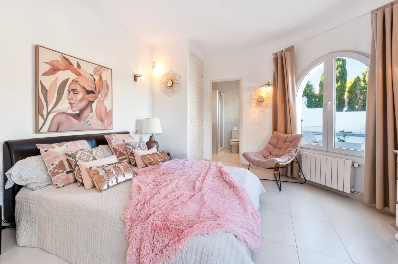 Bright bedroom featuring an en-suite bathroom, arched window overlooking the pool area, and a contemporary design with warm textiles and tiled flooring.