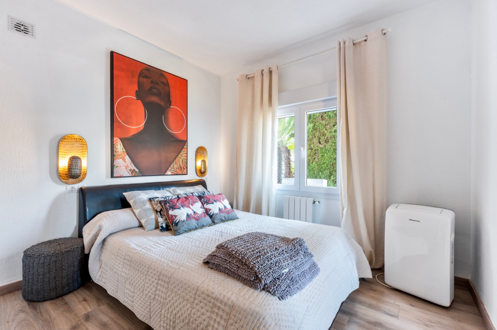 Bright bedroom featuring wood-effect flooring, a dark leather headboard, and vibrant wall art. Equipped with golden wall sconces, a radiator, and a portable AC unit.