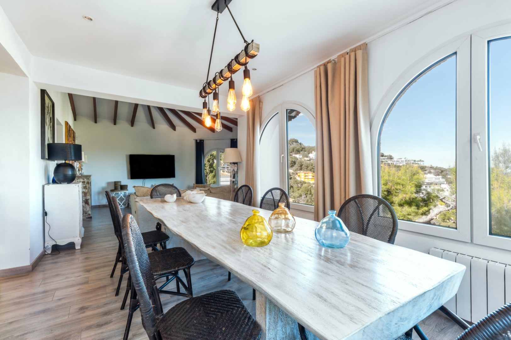 Bright dining area featuring a rustic wooden table, black wicker chairs, and arched windows with scenic views. Includes industrial-style lighting and exposed ceiling beams.