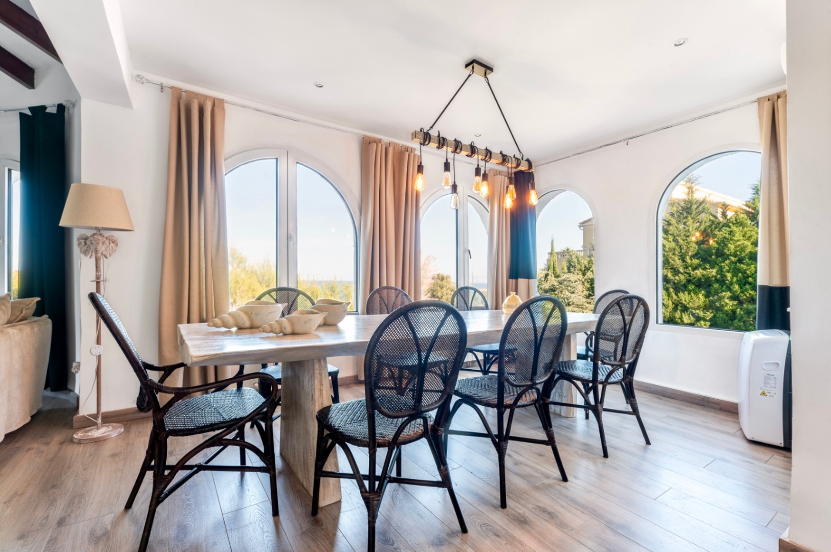 Bright dining room featuring a rustic wood table, black rattan chairs, and arched windows. An industrial beam pendant light adds a modern touch.