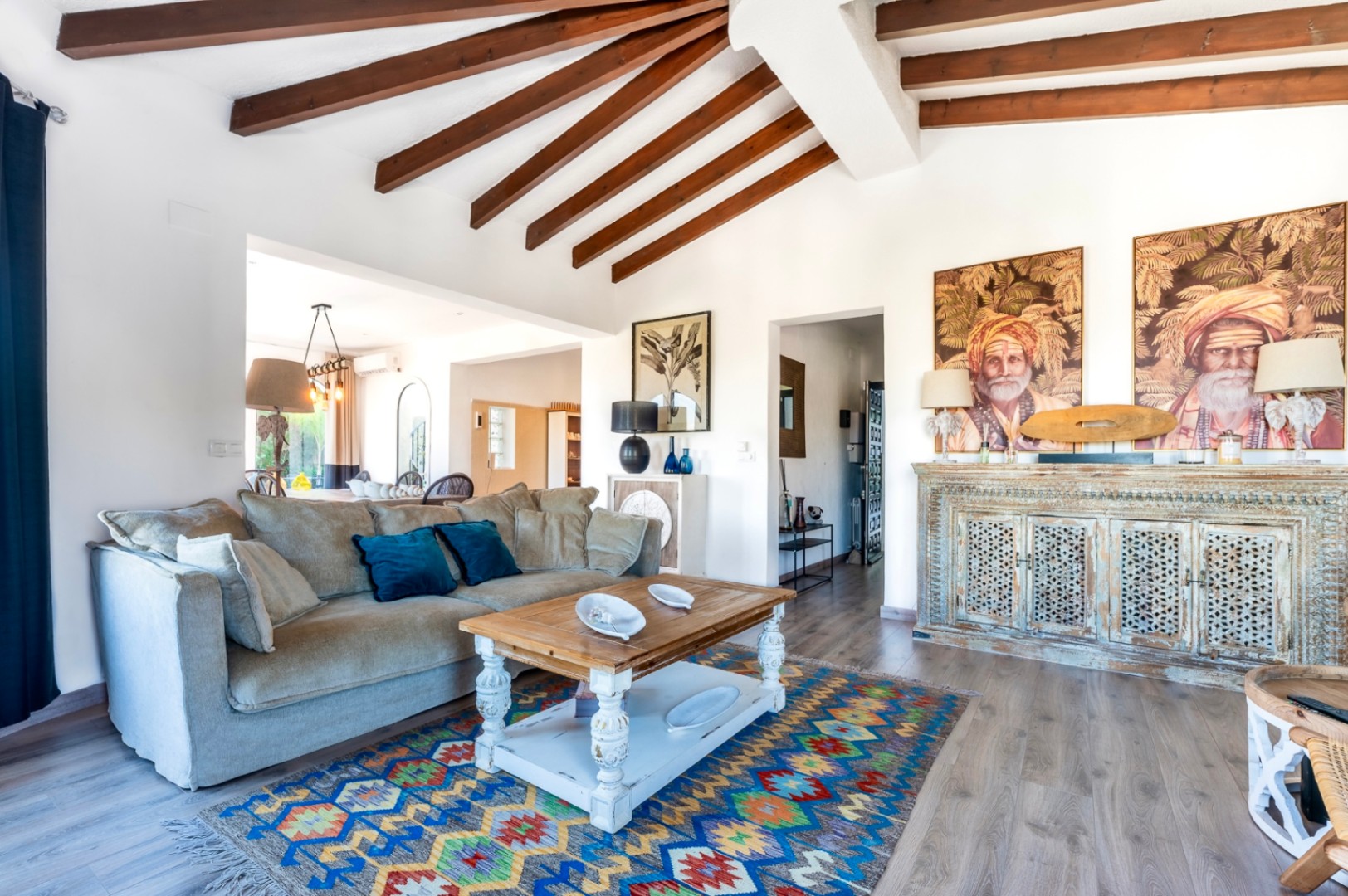 Bright living room featuring exposed dark wood ceiling beams, rustic furniture, and an ornate carved sideboard. The space includes wooden flooring and ethnic-inspired decor.