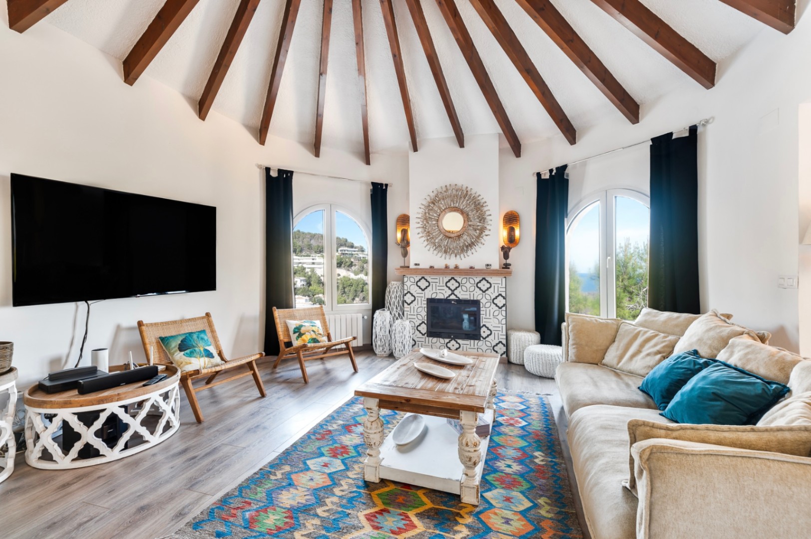 Circular living room featuring a vaulted ceiling with exposed wooden beams, a tiled fireplace, and arched windows offering views of the Mediterranean landscape.