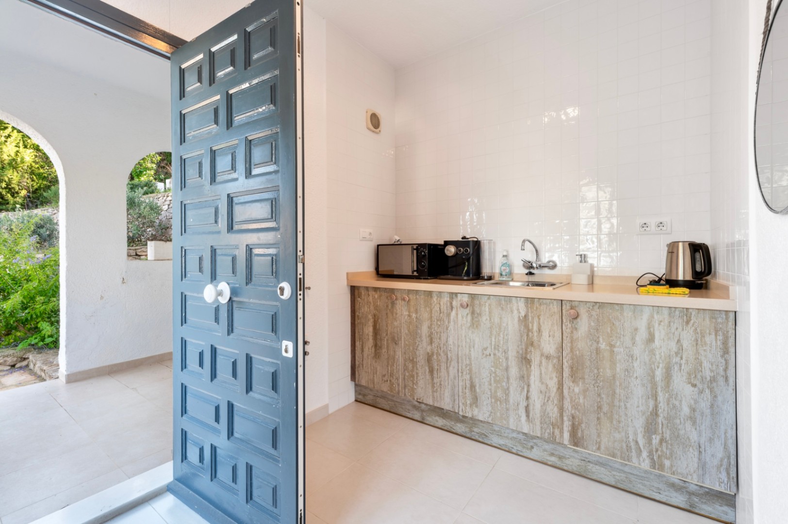Compact kitchenette featuring rustic wood-effect cabinetry and a light countertop, positioned next to a traditional blue paneled door leading to the terrace.