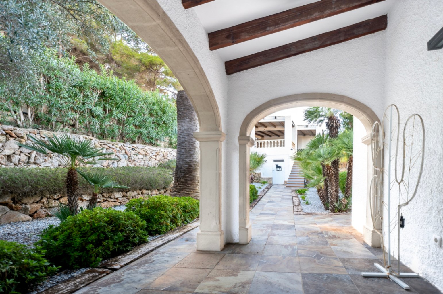 Covered terrace featuring traditional stone arches, exposed wooden beams, and tiled flooring, overlooking a landscaped garden with palm trees and stone walls.