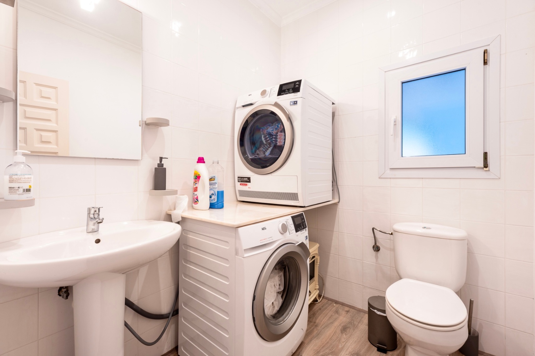 Functional laundry and guest bathroom featuring white tiled walls, AEG washer and dryer, a pedestal sink, and wood-effect flooring.