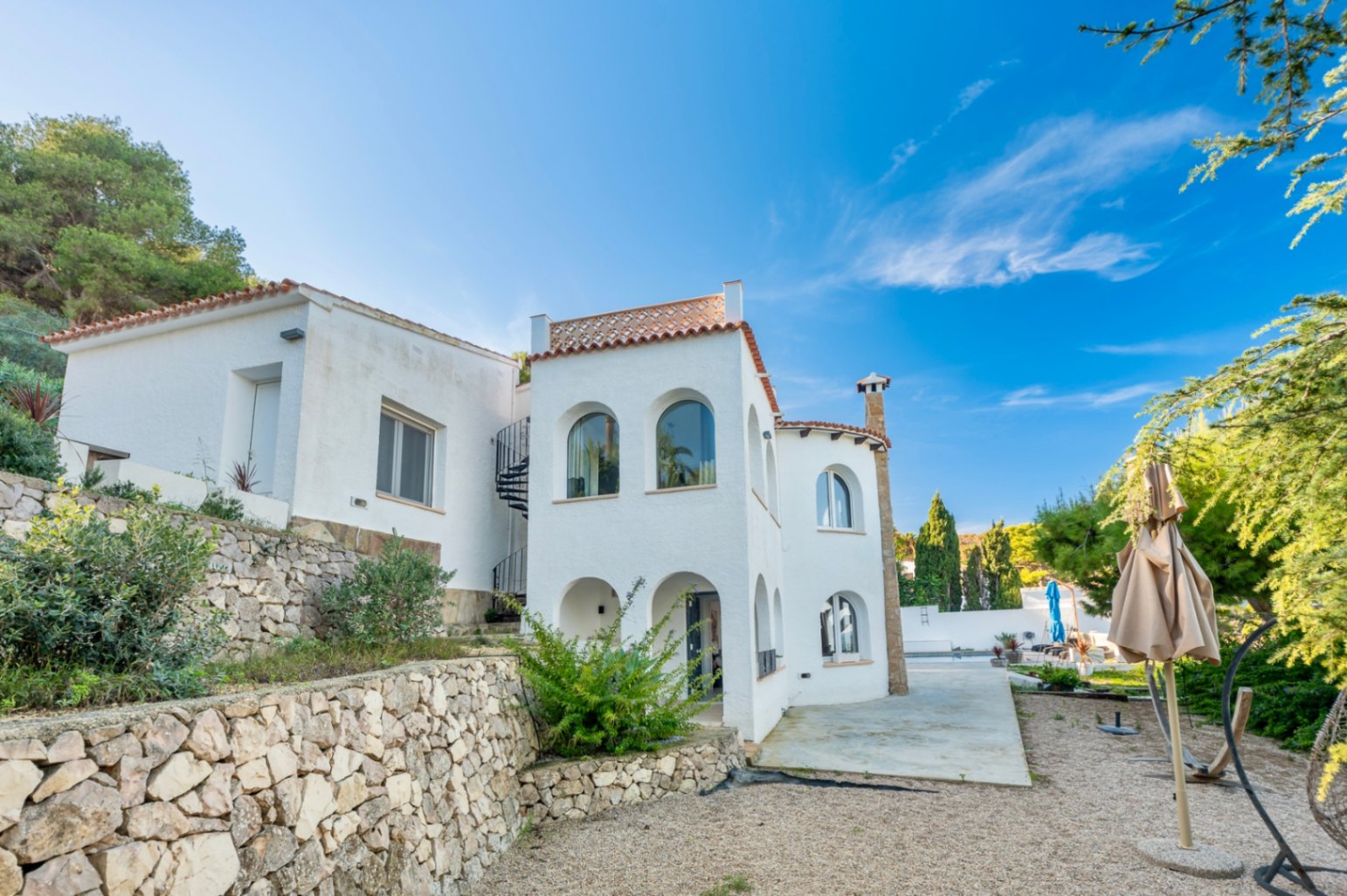 Mediterranean-style white facade featuring traditional arches, natural stone retaining walls, and an exterior spiral staircase set against a clear blue sky.