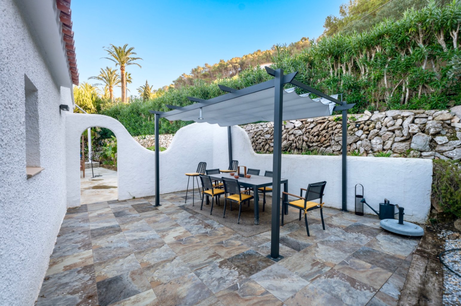 Outdoor dining area featuring a modern anthracite pergola, natural stone flooring, and white-washed walls set against a lush hillside.