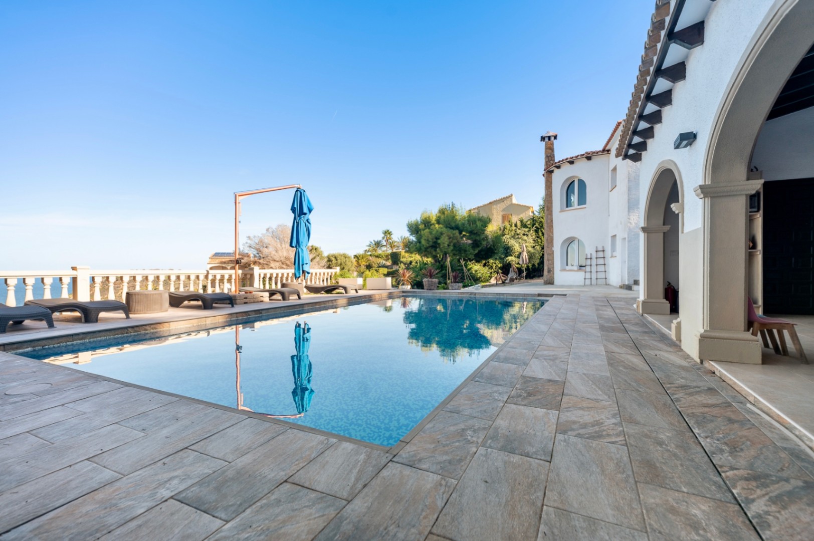 Outdoor terrace featuring a rectangular swimming pool, natural stone tiling, and sun loungers overlooking the Mediterranean coast and arched villa architecture.