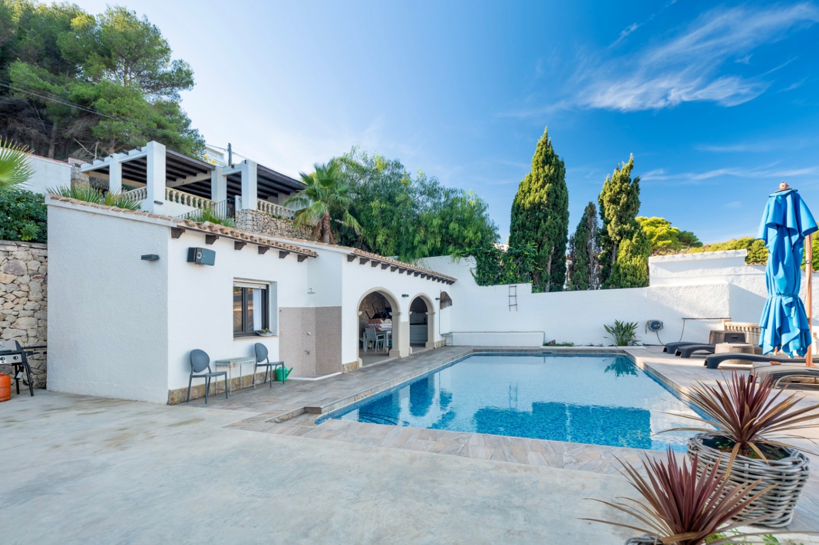 Rectangular swimming pool with tiled terrace, outdoor shower, and traditional arched porch. The private area features stone walls, cypress trees, and an upper pergola terrace.