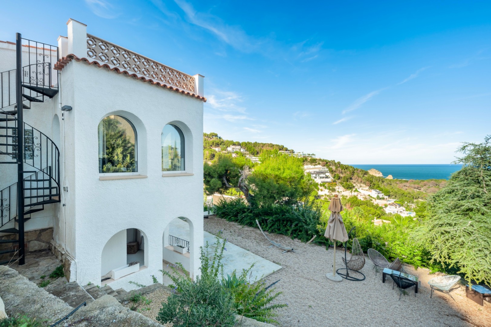 White villa facade featuring traditional arches, a black spiral staircase, and a gravel lounge area overlooking the Mediterranean Sea and coastal hills.