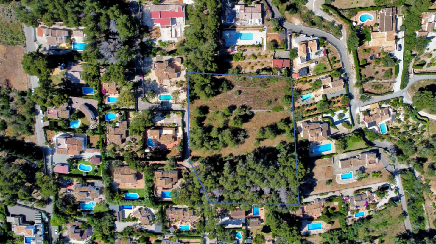 Aerial drone view of the large, flat plot outlined in blue, showing its natural pine vegetation and integration within an established residential neighborhood in Jávea.