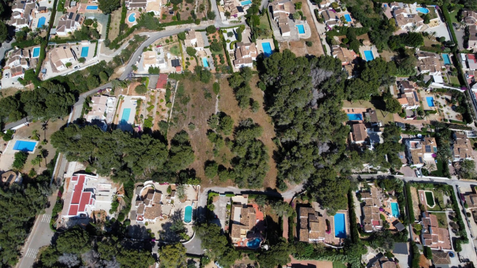 Aerial view of the expansive flat plot, situated within a developed residential area featuring villas with pools and mature pine trees.