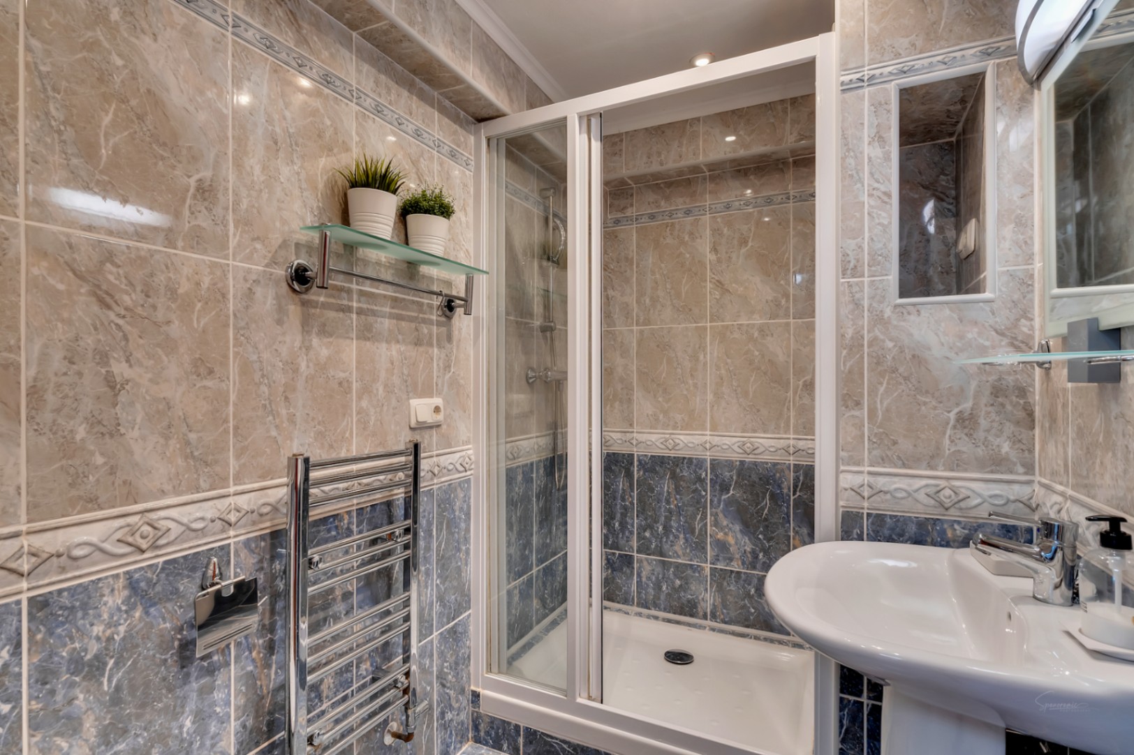 Bathroom featuring a walk-in shower with white-framed glass door, marble-effect tiling in beige and blue, and a chrome heated towel rail.