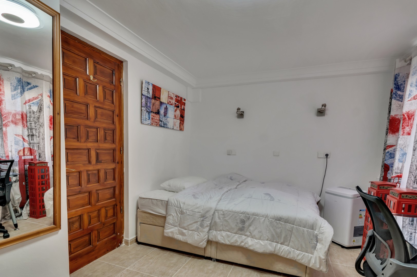 Bedroom featuring a double bed, ceramic tile flooring, and a traditional paneled wooden door. The room includes wall-mounted lighting and themed decor.