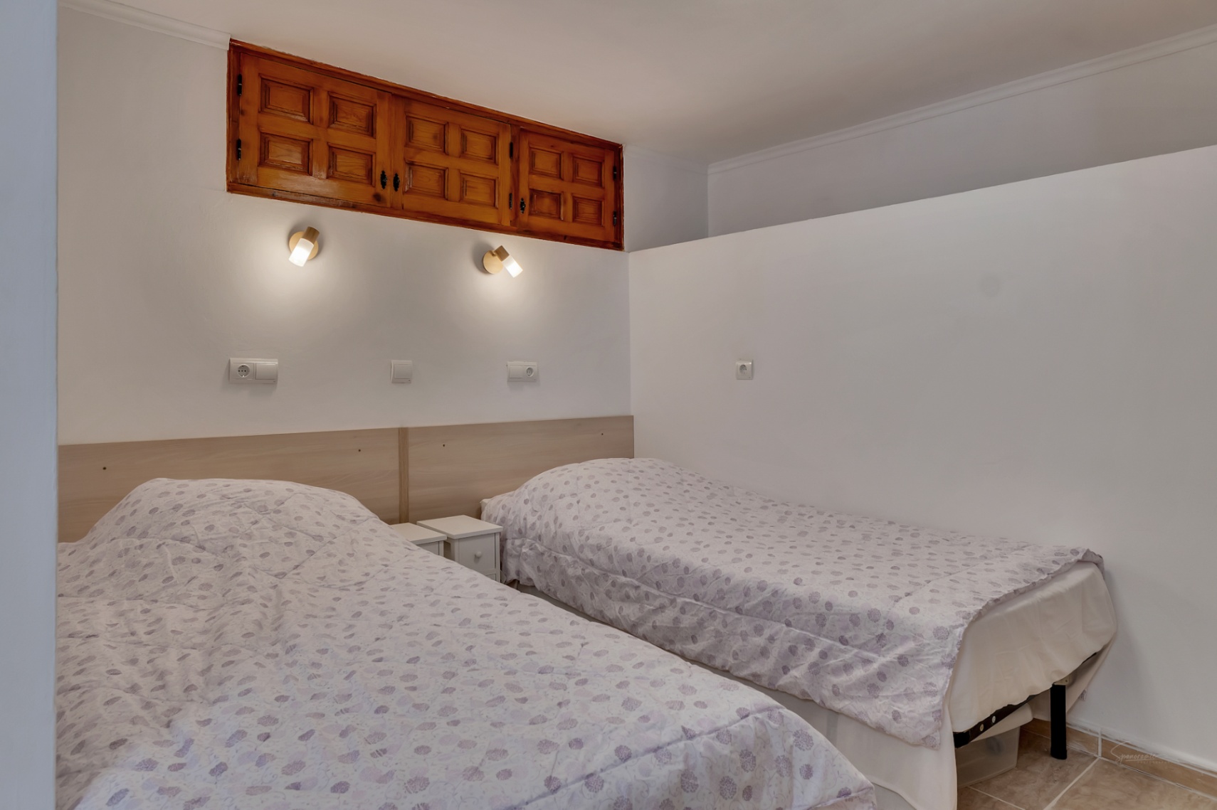 Bedroom featuring twin beds with a light wood headboard and wall-mounted reading lamps. A traditional wooden built-in cabinet is integrated into the wall above.
