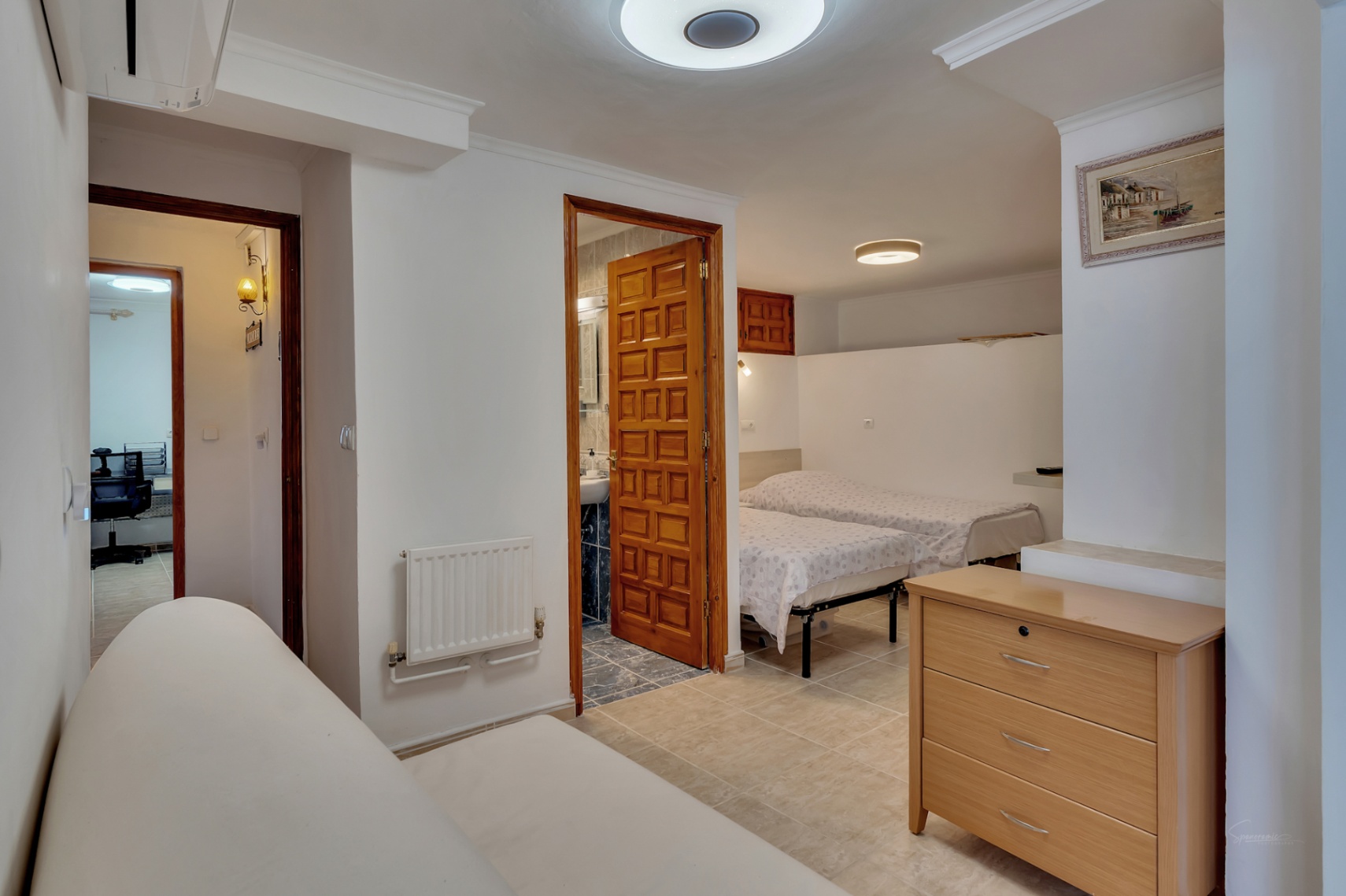 Bedroom with twin beds and en-suite bathroom featuring a carved wooden door. Includes wall radiator, ceramic flooring, and view towards an adjacent office space.
