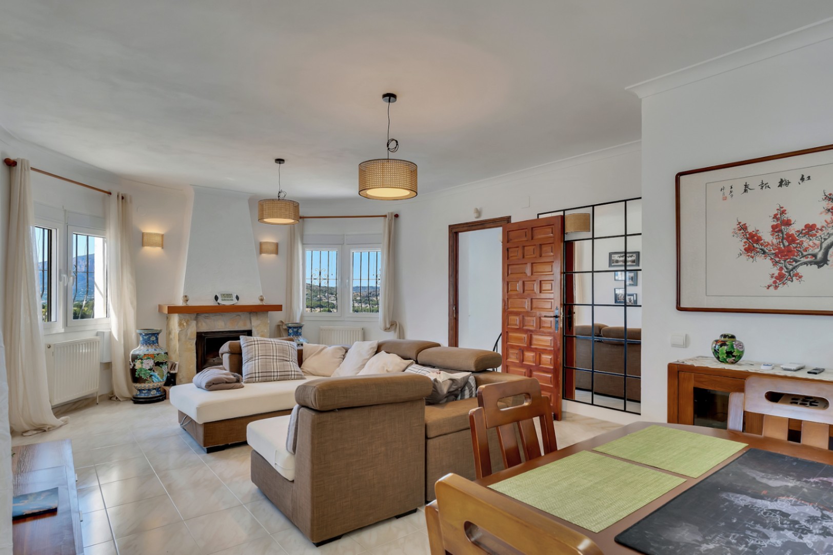 Bright living and dining area featuring a stone fireplace, modular seating, and views of the Montgó. Includes rattan pendant lighting, ceramic flooring, and a decorative grid mirror.