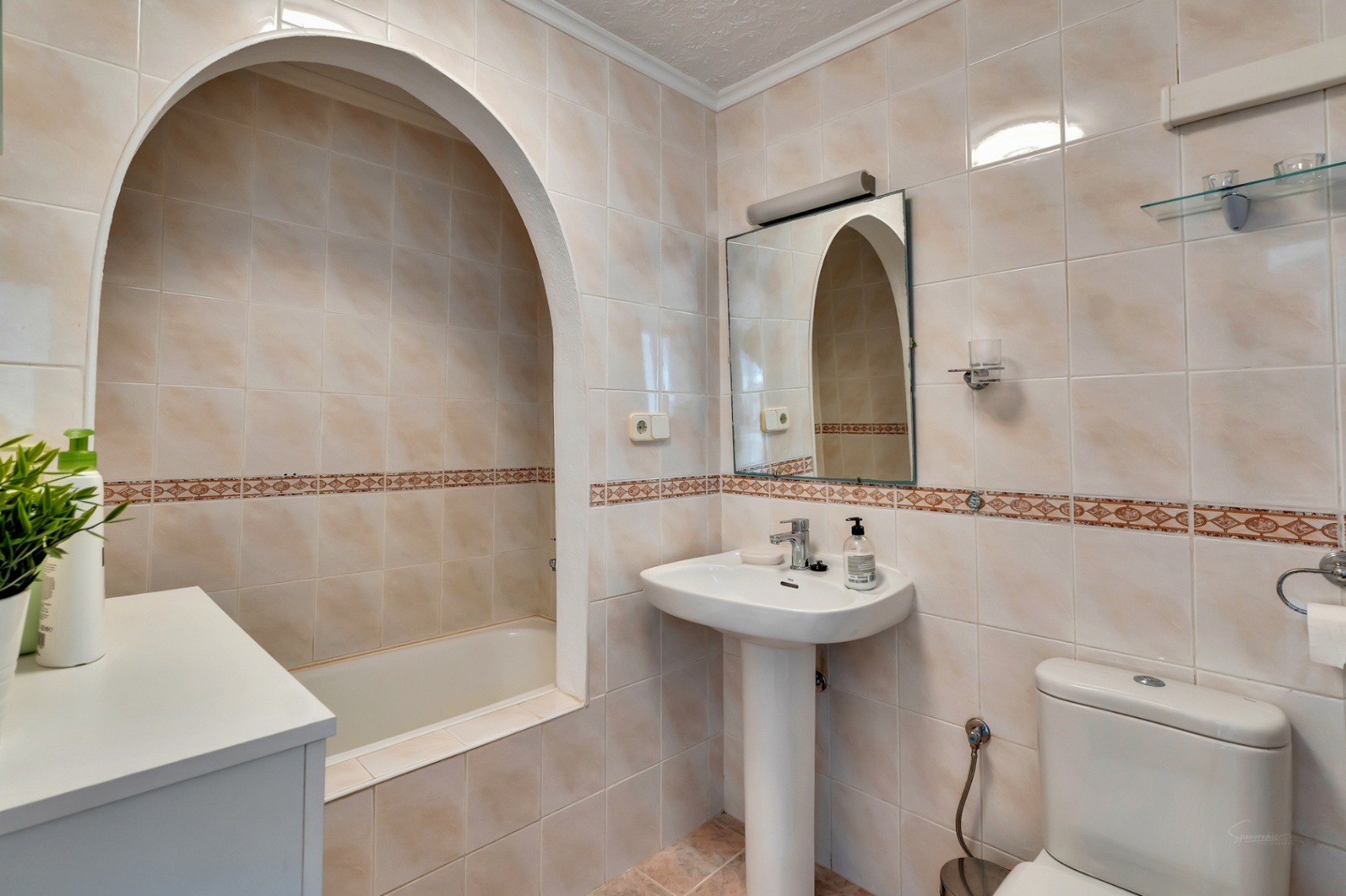 Classic bathroom featuring a bathtub set within a unique arched alcove, a pedestal sink, and cream-toned wall tiles with a decorative border.