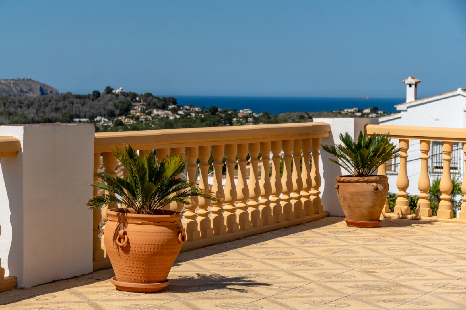 Elevated terrace featuring patterned floor tiles, a classic balustrade, and terracotta planters overlooking the Mediterranean Sea and coastal landscape.