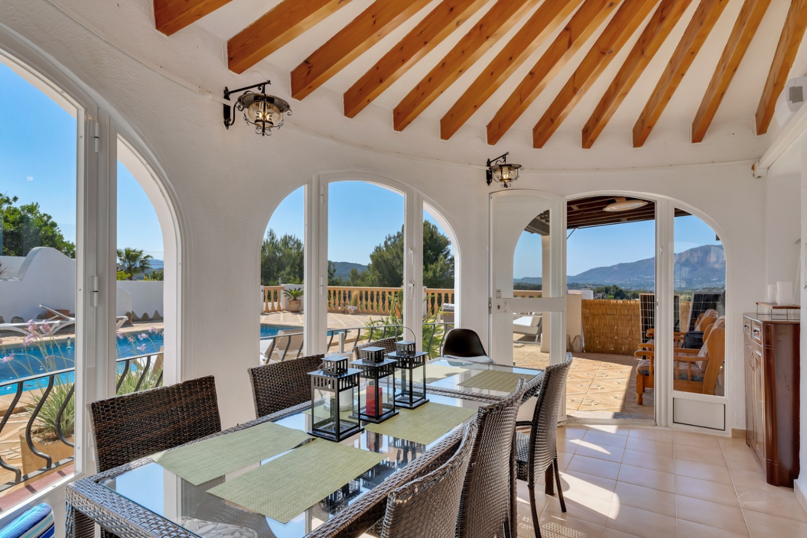 Enclosed dining area featuring radial wooden beams, arched windows overlooking the pool, and panoramic views of the Montgó mountain and landscape.