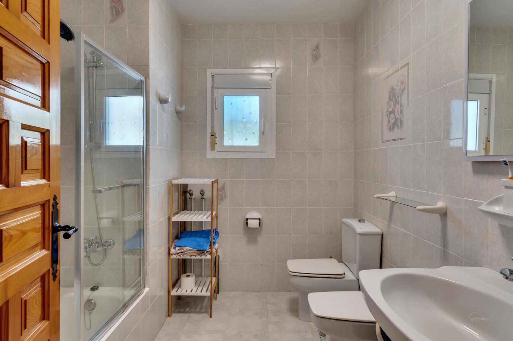 Functional bathroom featuring a bathtub with glass screen, white ceramic fixtures, wooden shelving, and tiled walls with subtle floral accents.