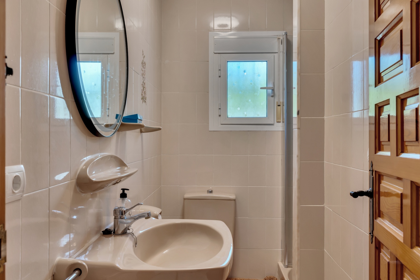 Functional bathroom featuring cream tiles, a round black-framed mirror, ceramic washbasin, and a small frosted window for natural light and ventilation.