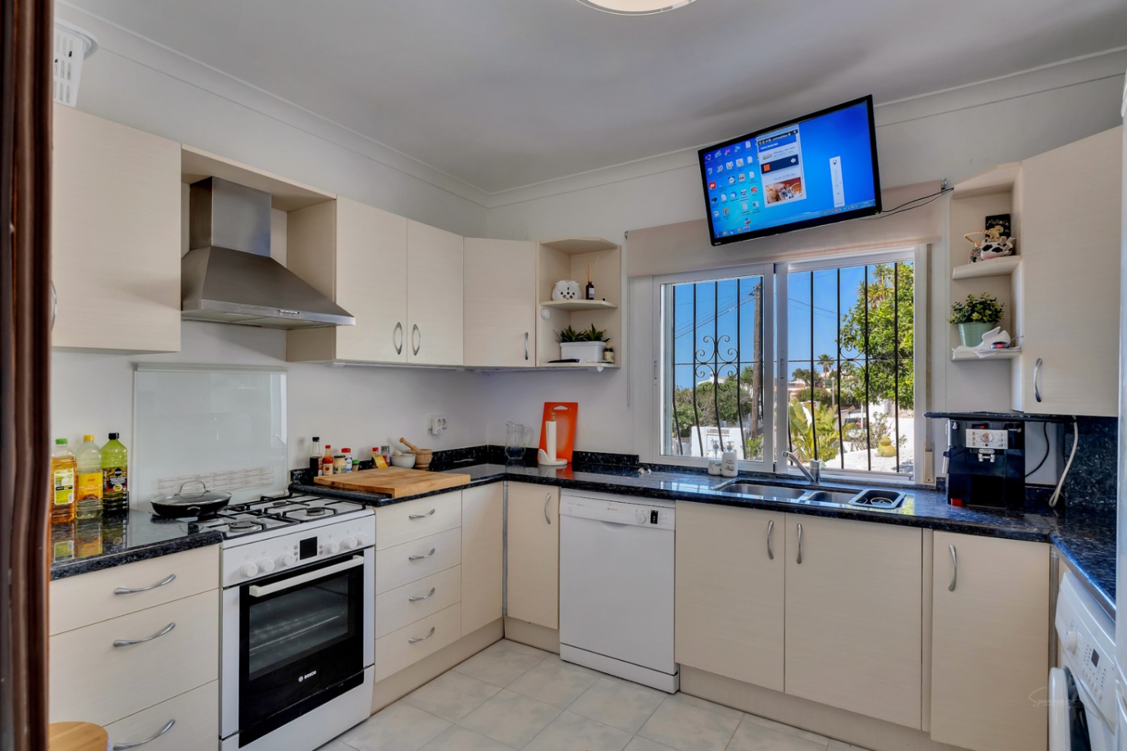 Functional L-shaped kitchen featuring dark granite countertops, cream cabinetry, integrated white appliances, and a window with garden views and a wall-mounted TV.
