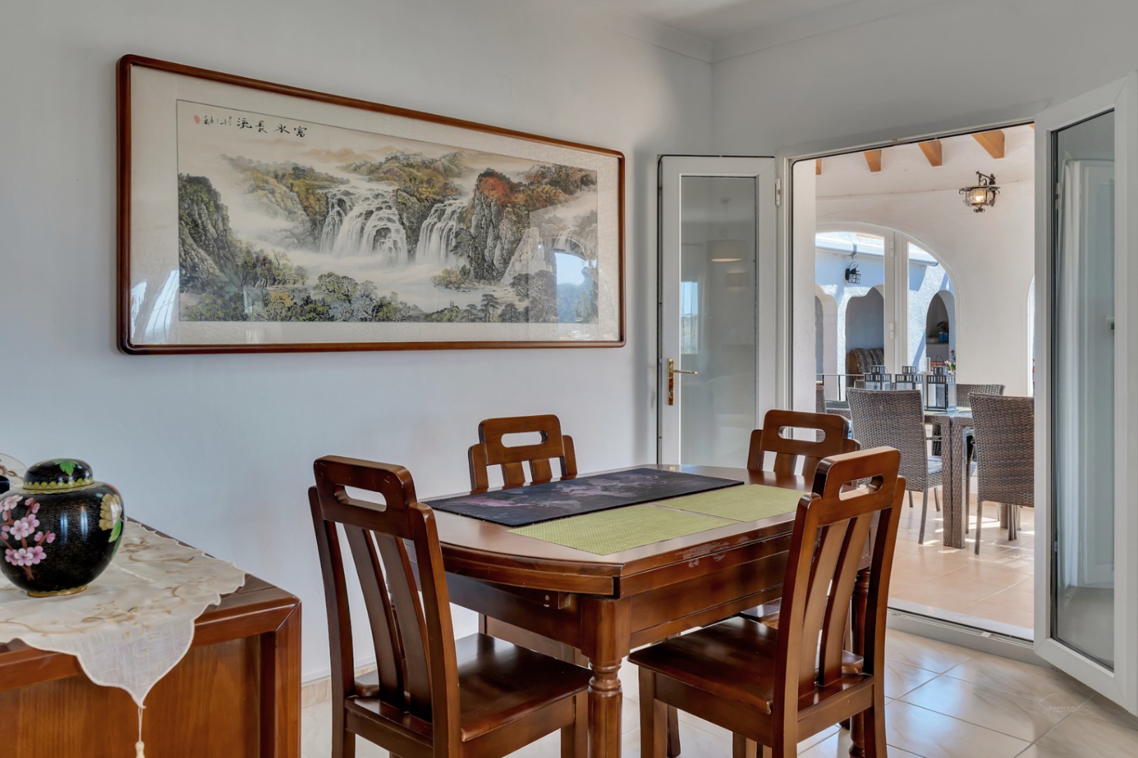 Indoor dining area featuring a wooden table set and a large landscape artwork. Glass doors open to a sunlit terrace with outdoor seating and arched details.
