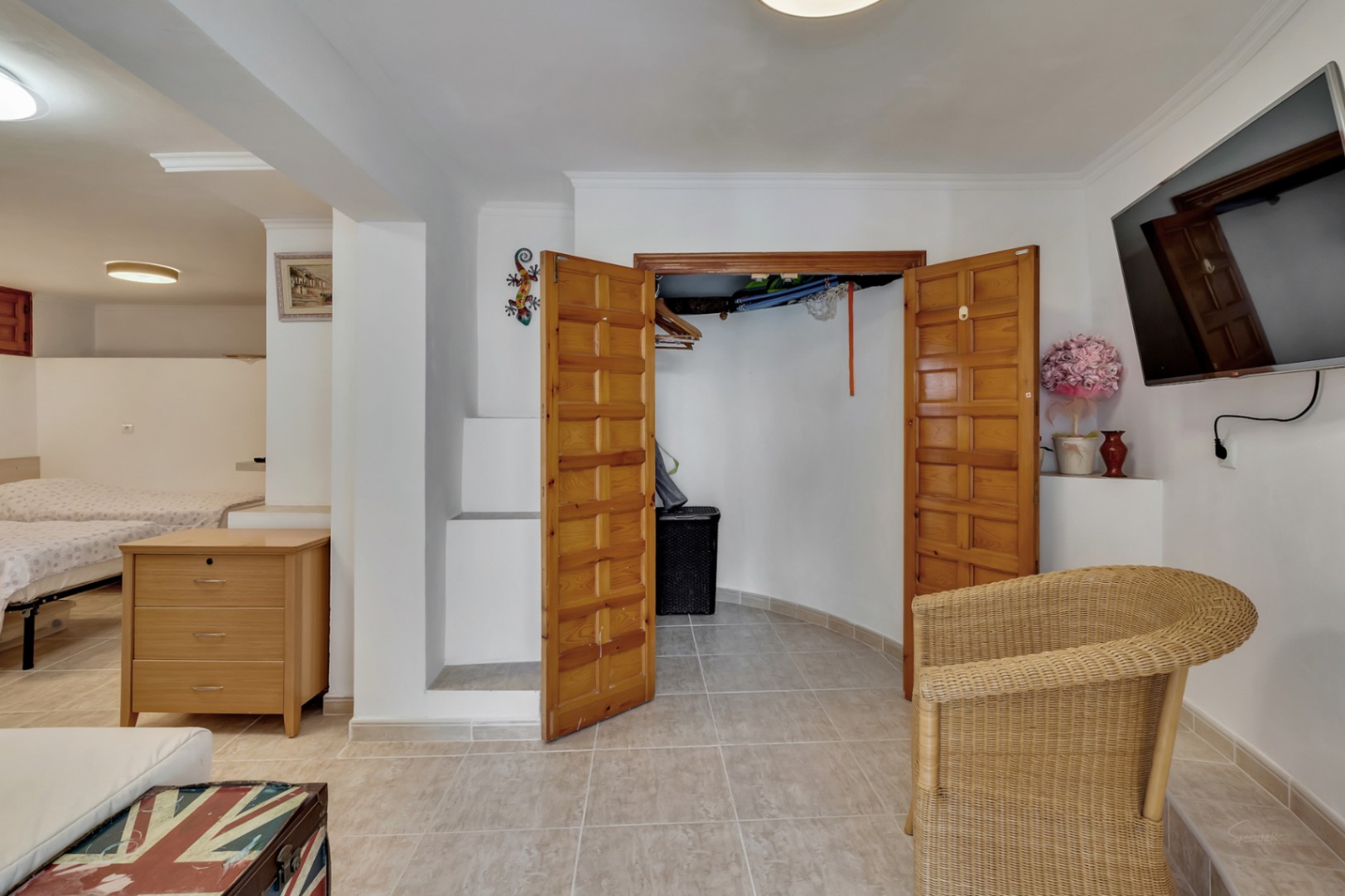 Lower floor bedroom featuring ceramic tiling, a built-in wooden closet, and a seating area with a wall-mounted TV and wicker armchair.