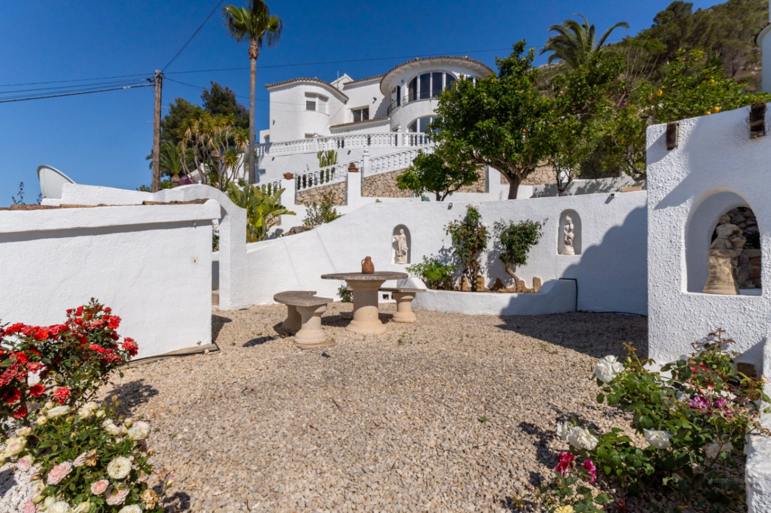 Mediterranean garden featuring a gravel patio, stone seating, and white-washed walls with decorative niches, set against the multi-level villa facade.