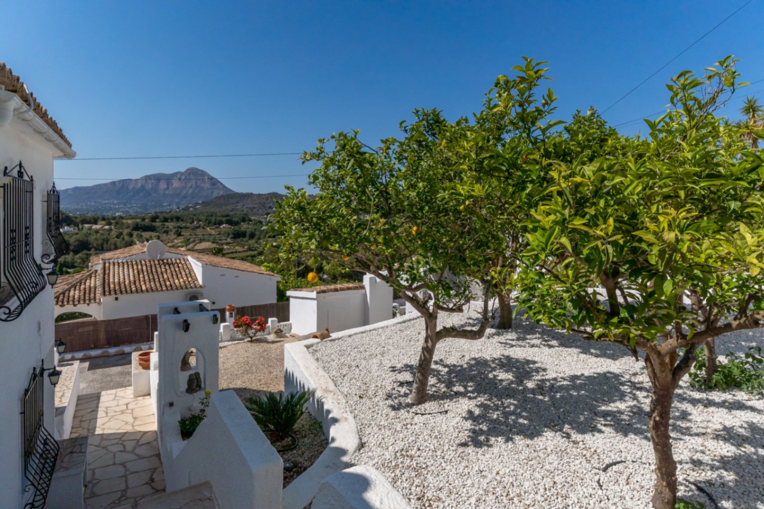 Mediterranean garden with white gravel, citrus trees, and clear views of the Montgó mountain. Natural stone pathway alongside the traditional white architecture.