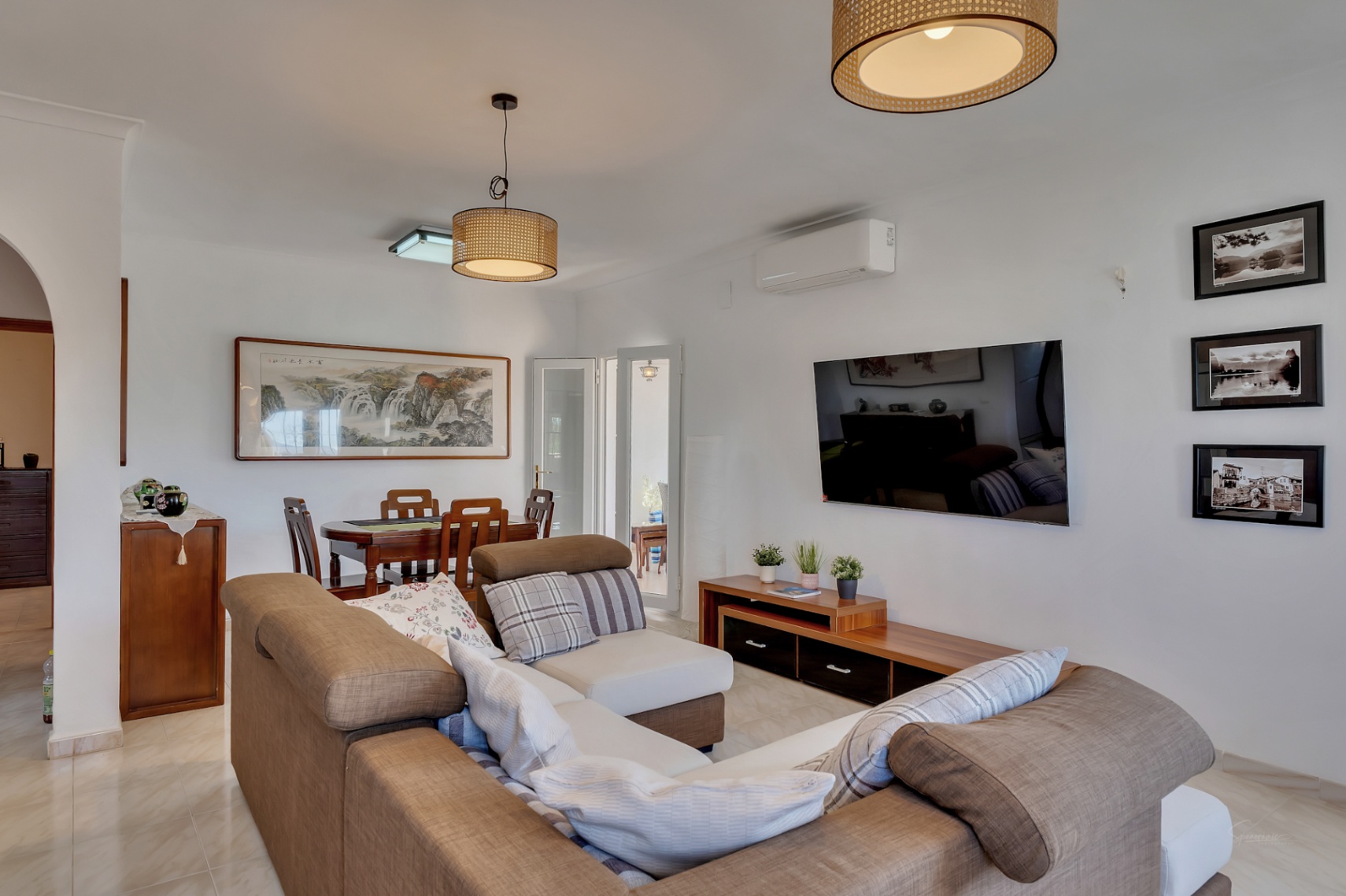 Open-plan living space featuring an L-shaped sofa, wooden dining set, and rattan pendant lights. The room includes air conditioning and neutral-toned tiled flooring.