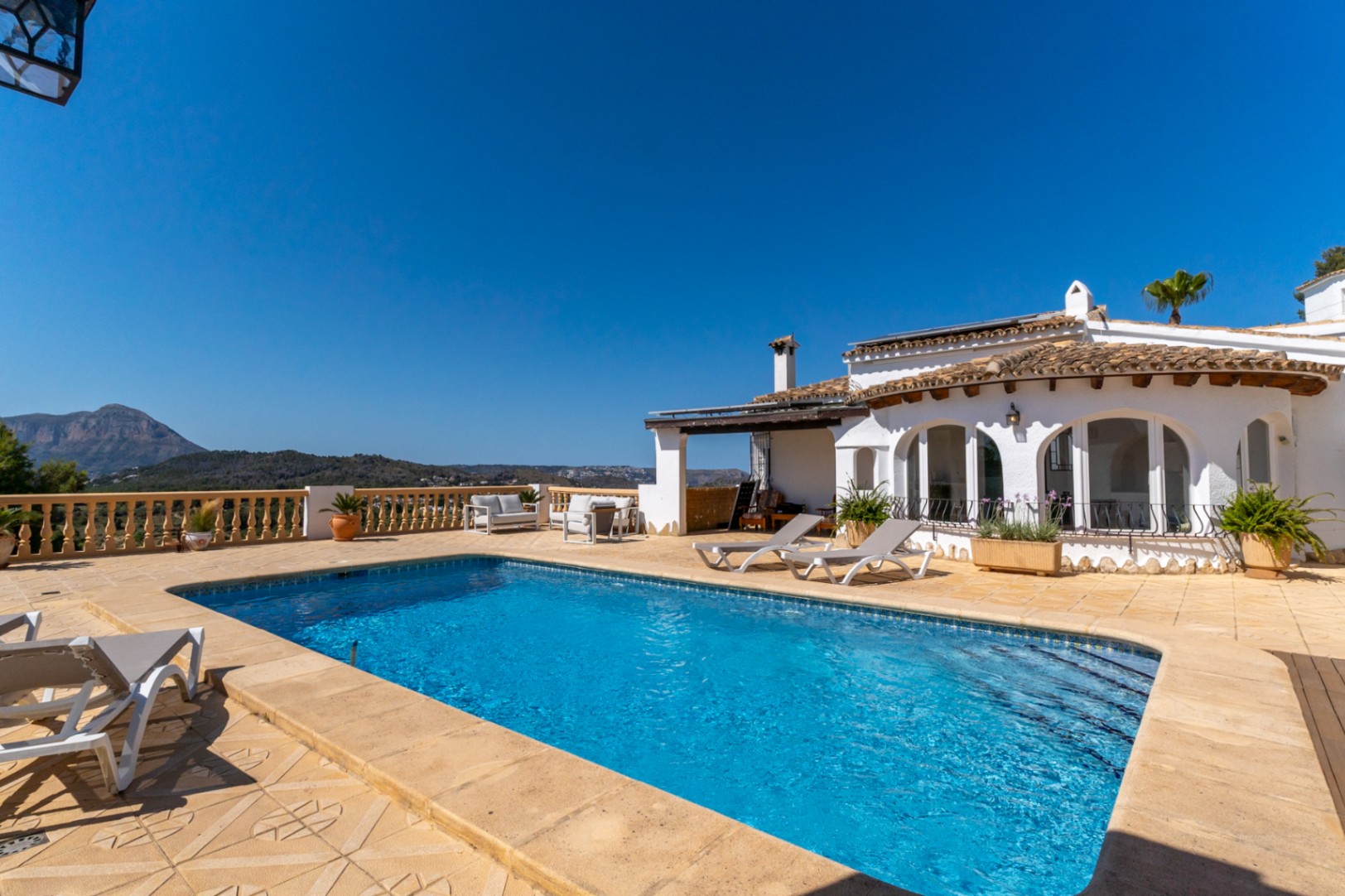 Outdoor swimming pool set within a spacious stone terrace offering panoramic views of the Montgó mountain. The white villa features traditional arches and rooftop solar panels.