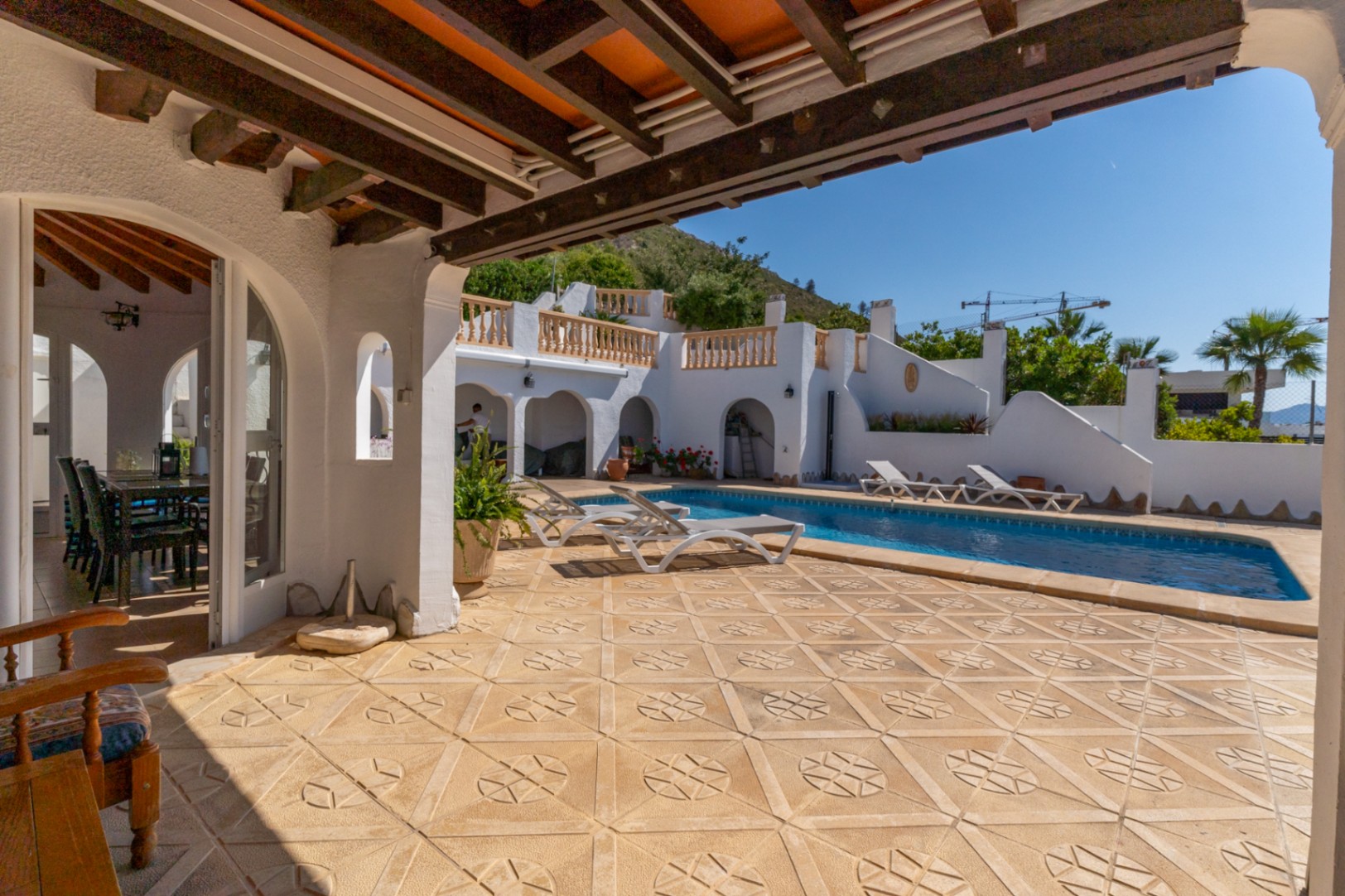 Outdoor terrace featuring patterned stone tiling, a rectangular swimming pool, and a covered porch with exposed wooden beams. The white facade overlooks the Montgó.