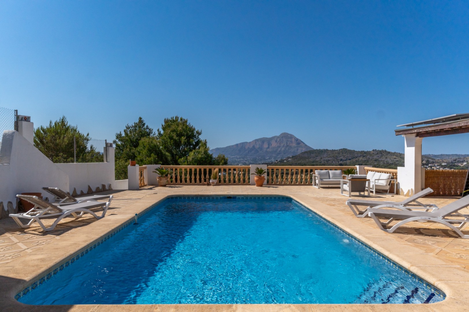 Rectangular swimming pool featuring a spacious stone terrace with sun loungers, outdoor seating, and panoramic views of the Montgó mountain.