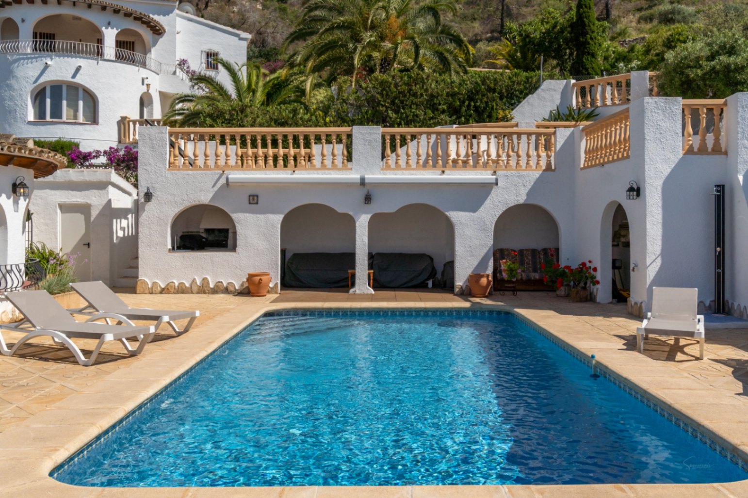 Rectangular swimming pool set in a stone terrace with sun loungers, featuring arched porch areas and traditional white balustrades against a lush backdrop.