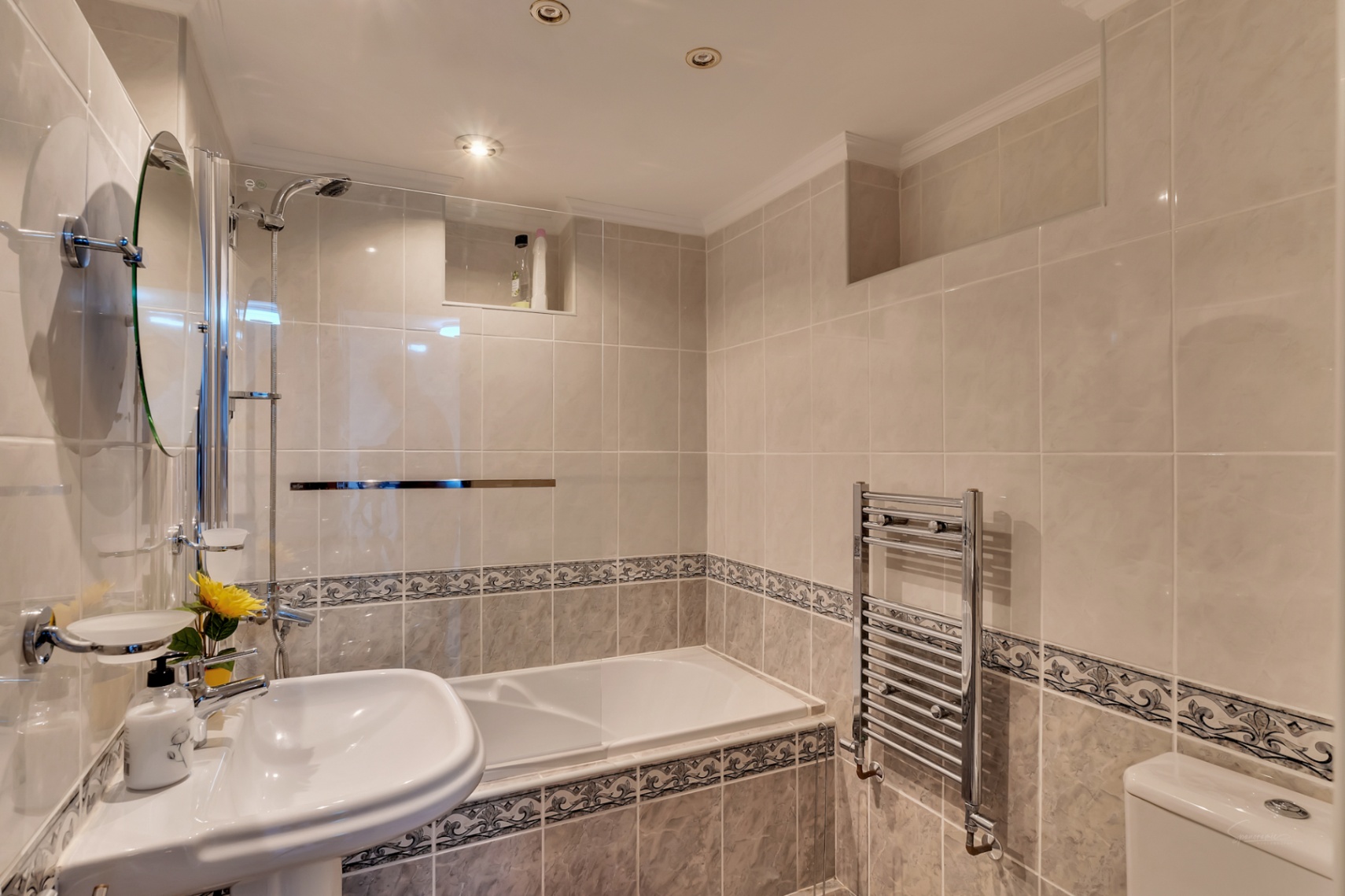 Tiled bathroom featuring a built-in bathtub with glass screen, pedestal sink, chrome towel radiator, and recessed ceiling spotlights.