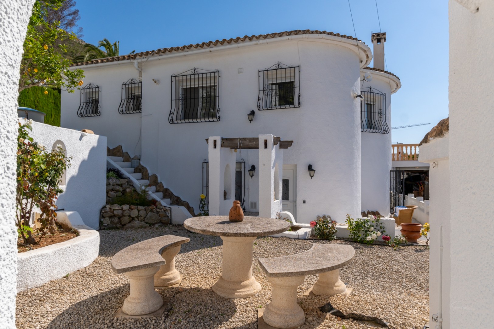 White Mediterranean villa exterior featuring decorative iron window grilles. A gravel seating area includes a circular stone table and curved benches.
