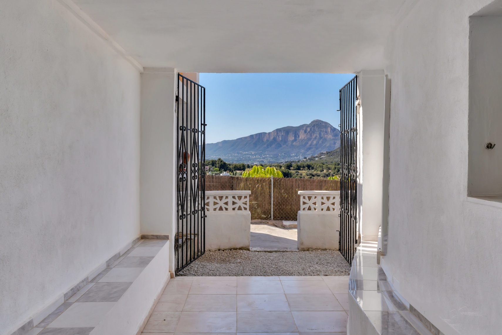 White-walled outdoor passage with tiled flooring, featuring an open wrought iron gate that frames panoramic views of the Montgó mountain.