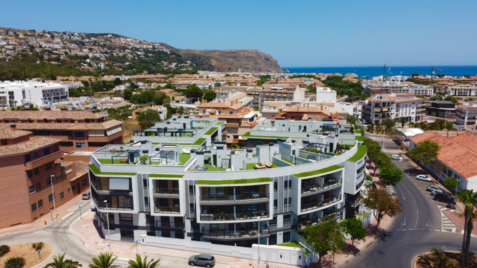 Aerial view of the modern residential complex featuring curved white architecture, private rooftop terraces with green finishes, and panoramic views towards the Mediterranean coast.