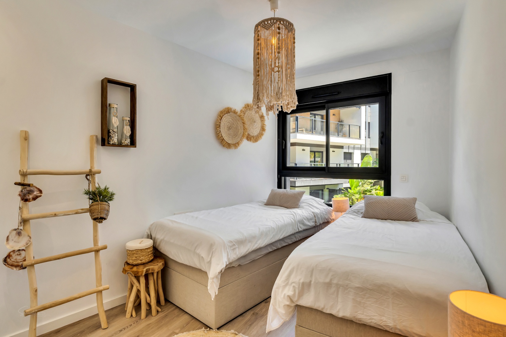 Bedroom featuring twin beds and boho-chic decor, including a macramé pendant light and wooden accents, with views through a large black-framed window.