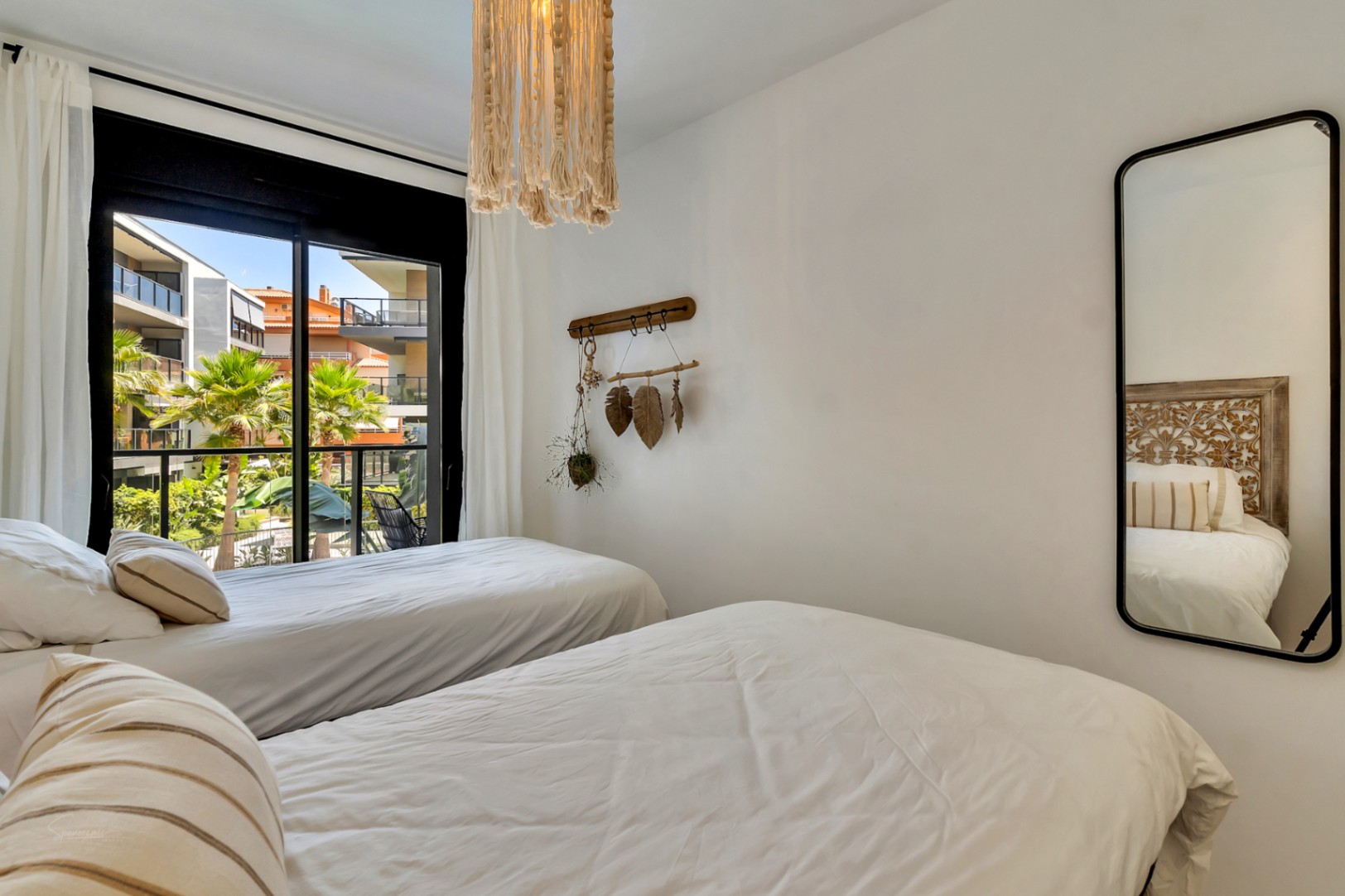 Bedroom with twin beds, a macramé pendant light, and a vertical mirror. Large sliding glass doors provide views of the complex's gardens and palm trees.