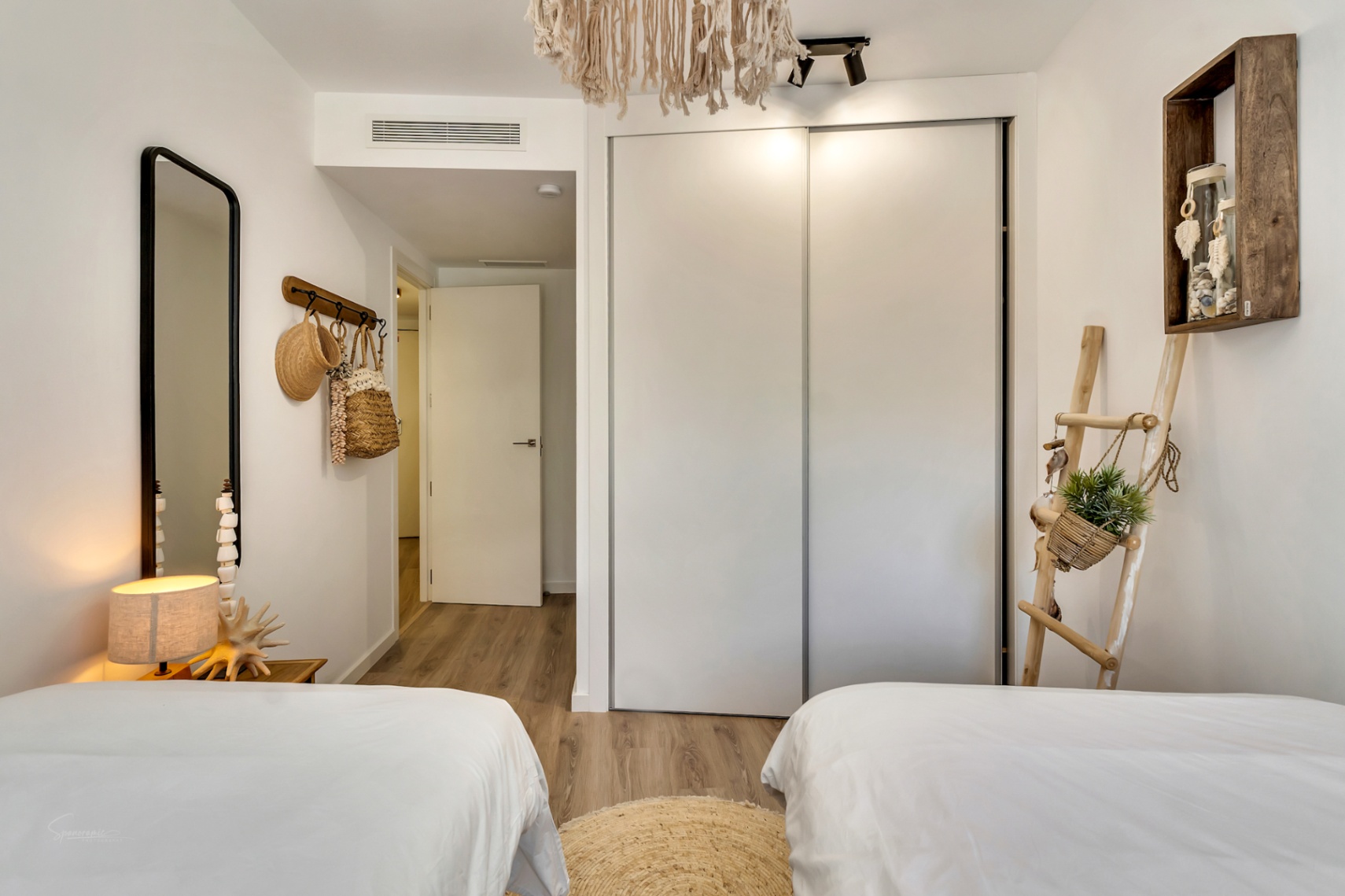 Bright bedroom featuring twin beds, integrated sliding-door wardrobes, light wood flooring, and Mediterranean-inspired decor with natural fiber accents.