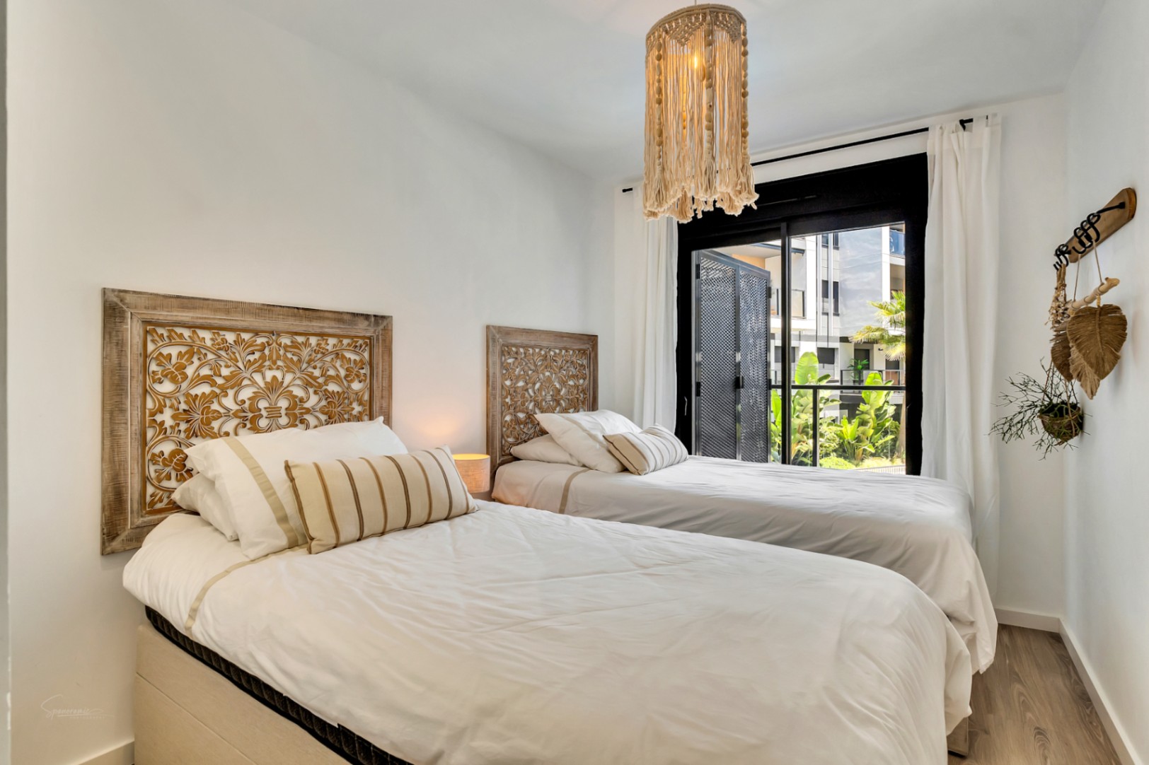 Bright bedroom featuring twin beds with ornate carved wooden headboards and a macramé pendant light. Large sliding doors open to a terrace with lush garden views.