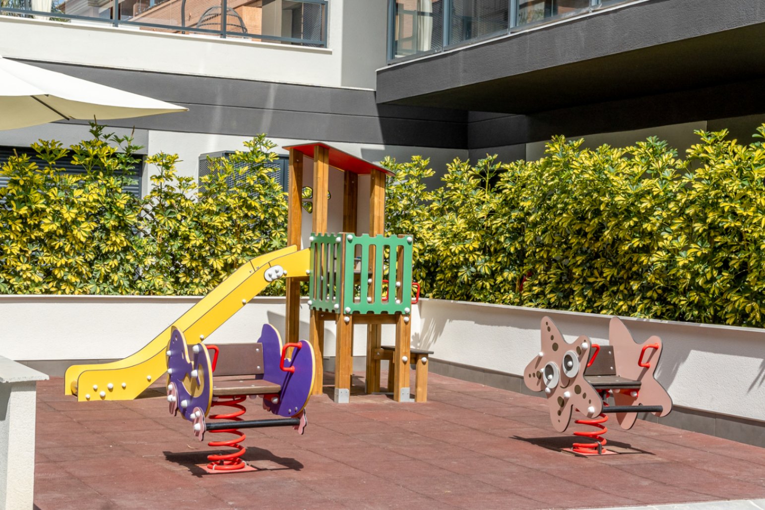 Communal children's playground featuring a safety rubber floor, yellow slide, and spring riders, set against manicured hedges and modern residential architecture.