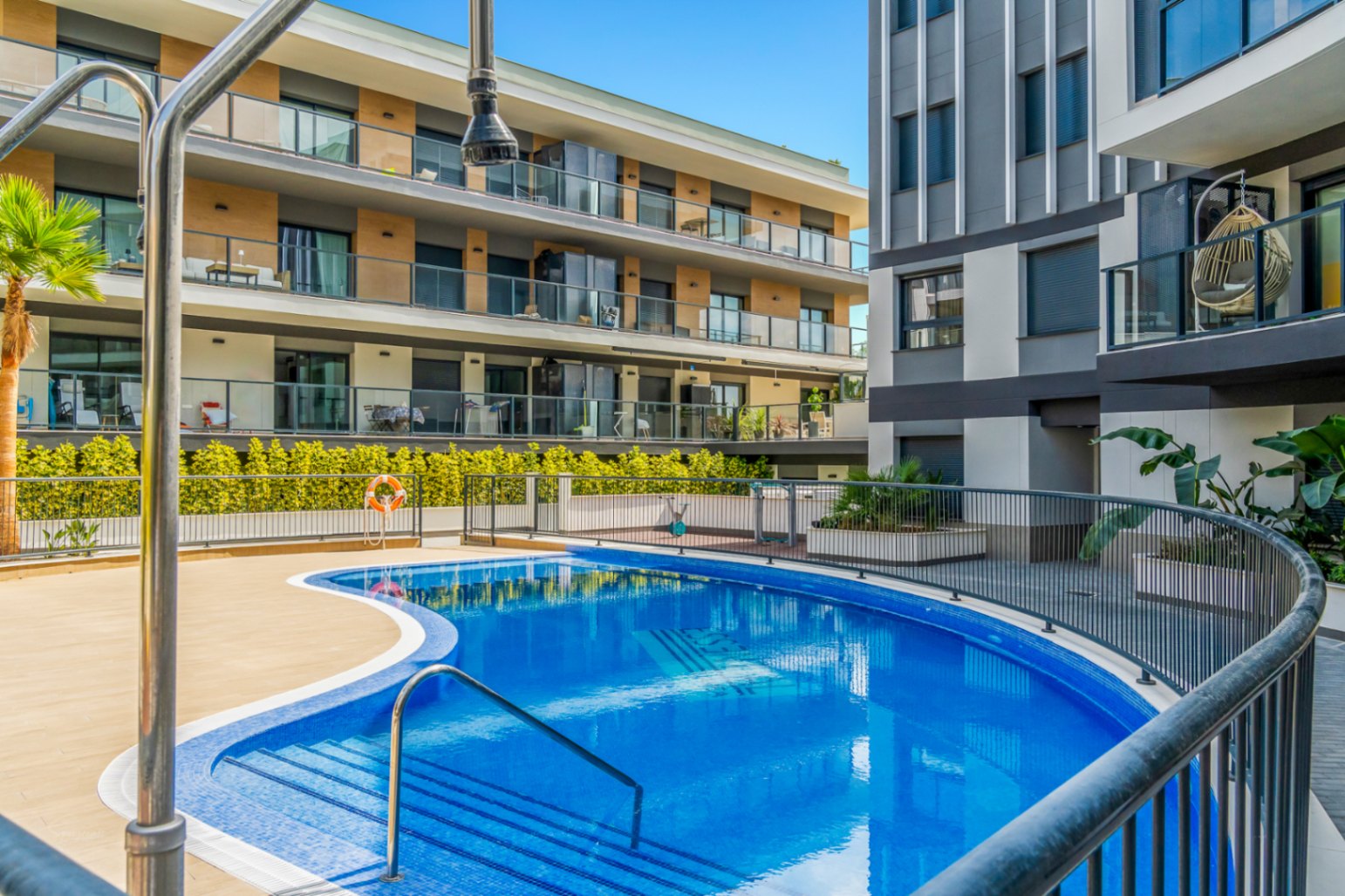 Communal curved swimming pool featuring blue mosaic tiling, a sun terrace, and safety railings, set against the modern architectural backdrop of the 2023 complex.
