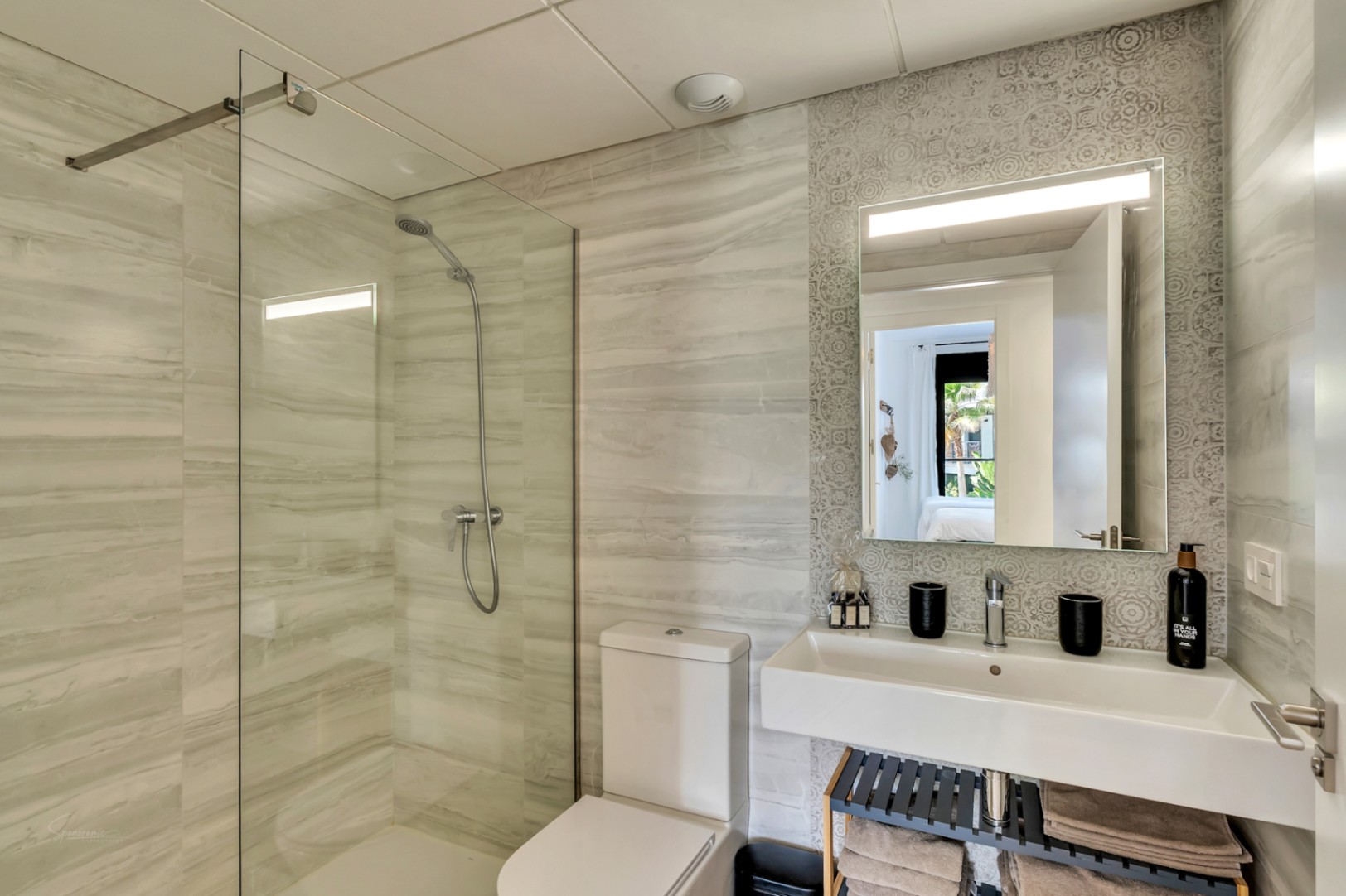 Modern bathroom featuring a walk-in shower with glass partition and marble-effect tiling. A floating washbasin sits against a patterned accent wall with an illuminated mirror.